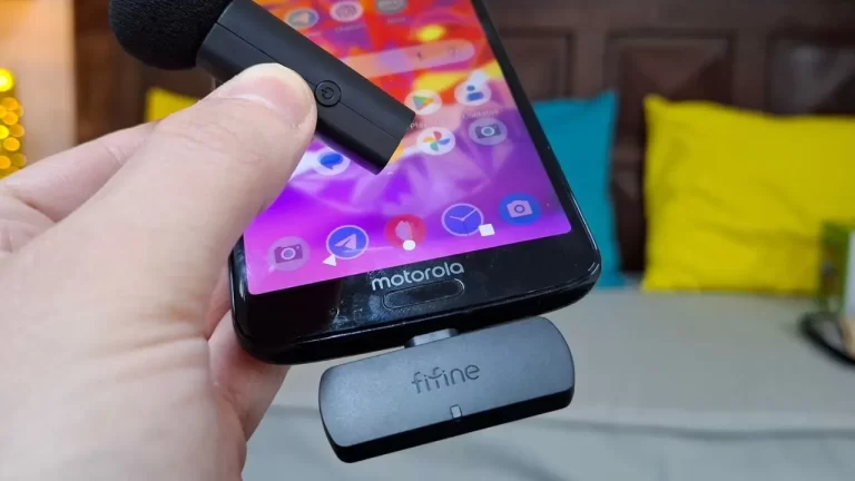 Mão segurando um microfone de lapela sem fio próximo a um celular Motorola que possui um receptor Fifine conectado na porta USB-C.