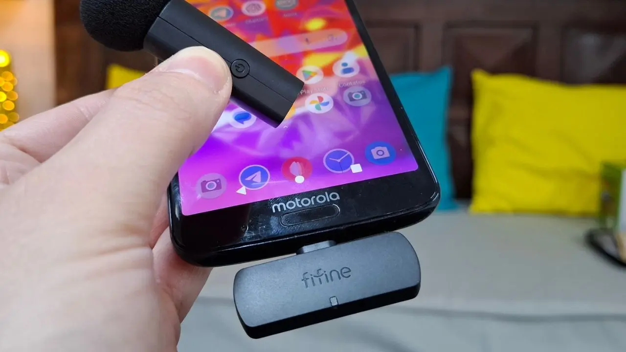Mão segurando um microfone de lapela sem fio próximo a um celular Motorola que possui um receptor Fifine conectado na porta USB-C.