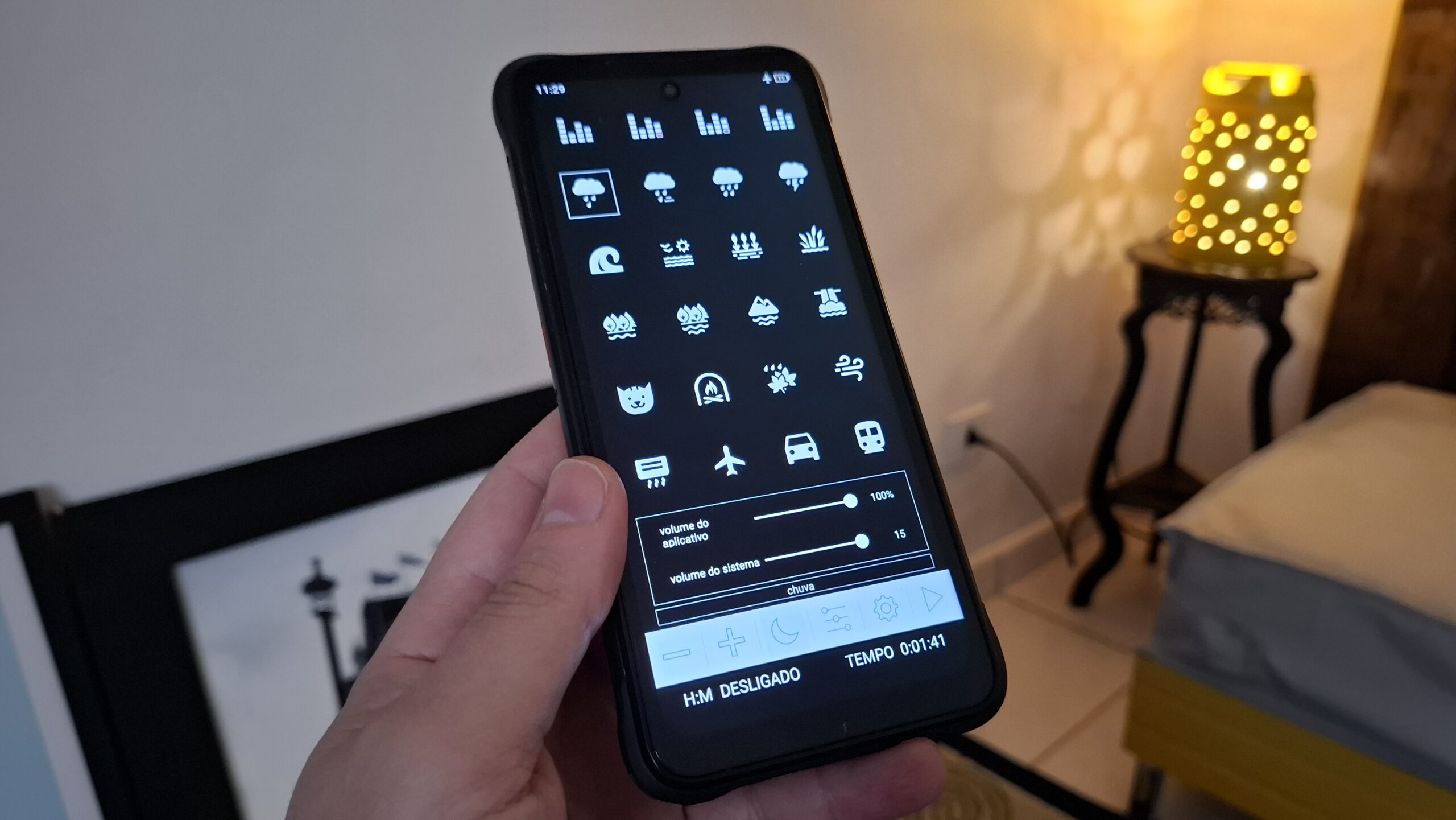 Mão segurando um smartphone antigo em um ambiente escuro, exibindo a interface do aplicativo de ruído branco com diversos ícones de sons relaxantes.