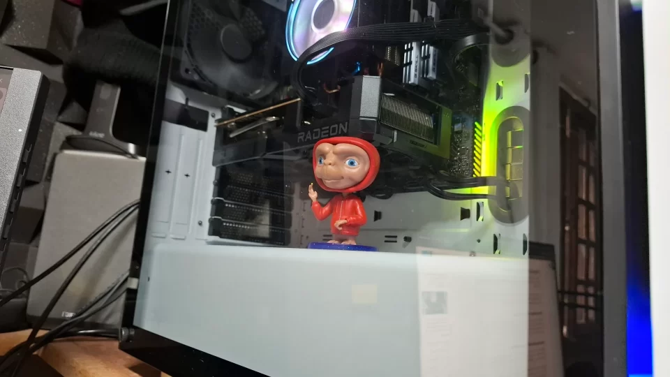 Boneco do personagem E.T. utilizado como suporte criativo para evitar a inclinação de uma placa de vídeo Radeon RX 7600 TUF dentro de um gabinete gamer com iluminação RGB.