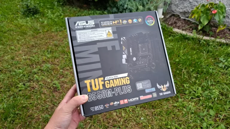 Mãos segurando a caixa da placa-mãe ASUS TUF Gaming B550M-Plus em um ambiente externo com grama ao fundo, mostrando a embalagem do produto.
