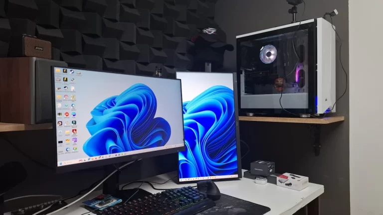 Foto de um setup gamer de alto desempenho com dois monitores mostrando o Windows 11 e um gabinete branco com iluminação RGB, em um ambiente com isolamento acústico.