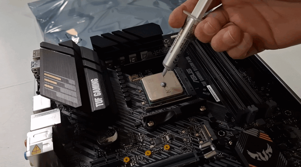 Mão aplicando uma pequena gota de pasta térmica cinza sobre o centro de um processador de computador instalado em uma placa-mãe.