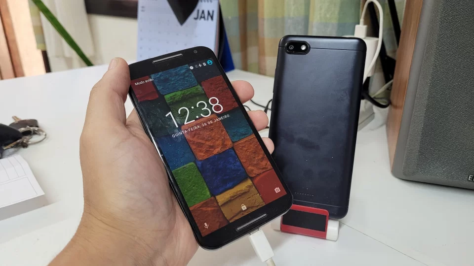 Mão segurando um smartphone antigo ligado ao lado de outro aparelho desativado sobre uma mesa de escritório.