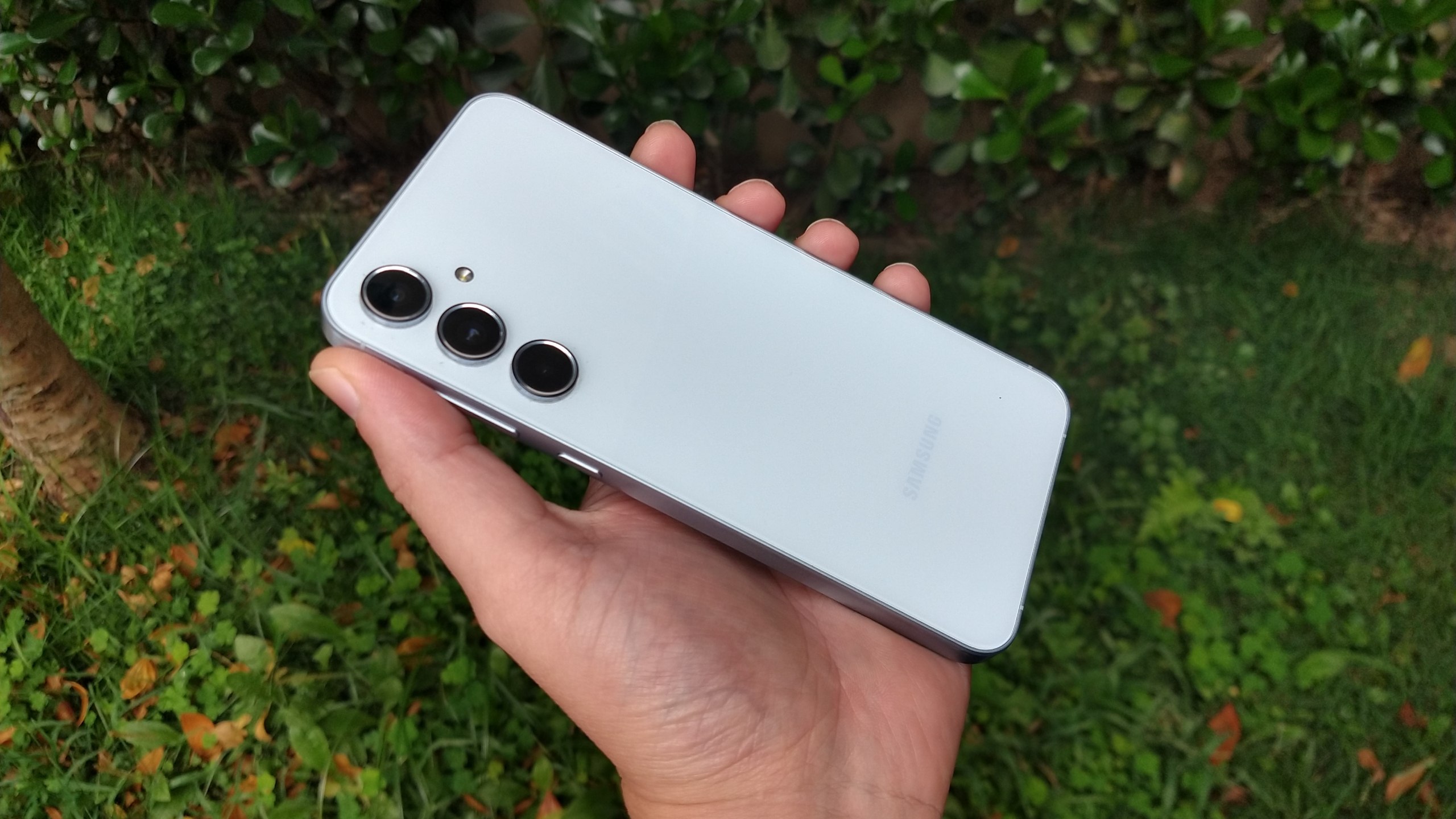 Mão segurando um smartphone Samsung Galaxy A55 de cor clara em um ambiente externo com gramado ao fundo.