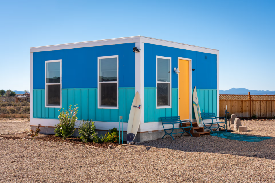 Vista externa da casa modular Boxabl Casita Studio na cor azul, com porta laranja e várias janelas, instalada em um ambiente desértico com montanhas ao fundo.
