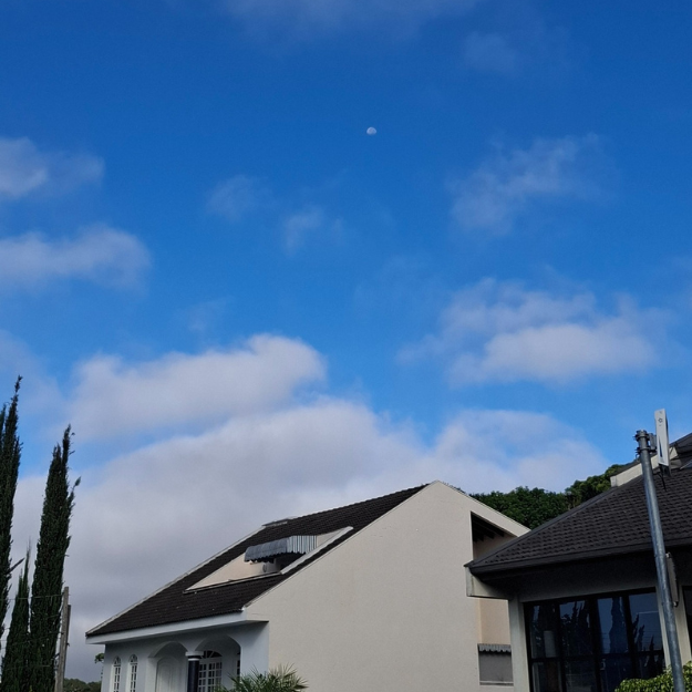 Fotografia de um céu azul vibrante com nuvens brancas esparsas, onde se vê a Lua diurna em fase minguante no centro, posicionada acima de casas residenciais.
