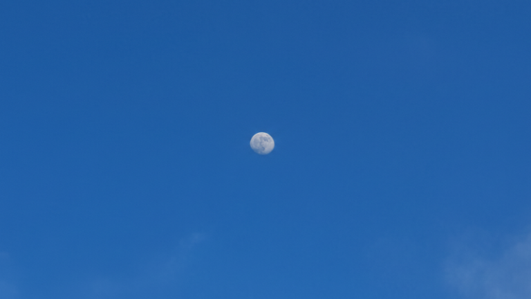 Fotografia de um céu azul vibrante com nuvens brancas esparsas, onde se vê a Lua diurna em fase minguante no centro.