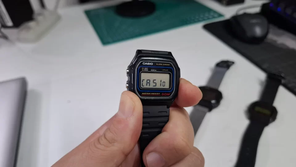 Close em um relógio Casio W-59 exibindo a palavra "CASio" no visor digital após pressionar o botão lateral.