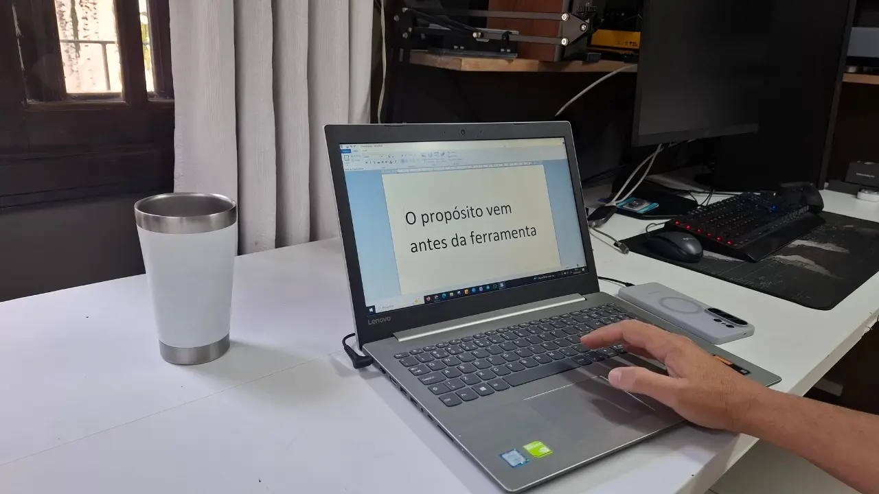 Visão em primeira pessoa de um ambiente de home office organizado. Uma mão opera o touchpad de um notebook cinza sobre uma mesa branca. Na tela do notebook, um documento de texto está sendo editado. Ao lado, um copo térmico branco. O ambiente é bem iluminado, sugerindo um espaço de trabalho focado e profissional para criação de conteúdo com IA.