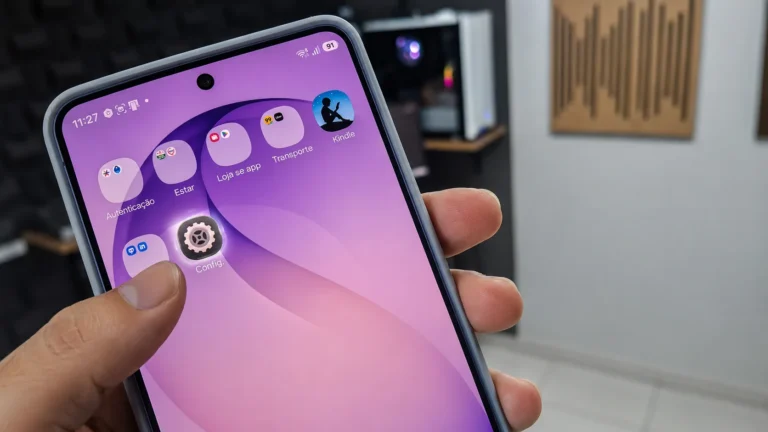 Mão segurando um smartphone Android com a tela inicial aberta, exibindo aplicativos e um fundo lilás, em um cenário de estúdio de gravação de tecnologia.