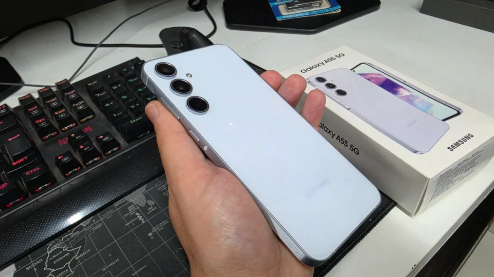 Mão segurando um smartphone Samsung Galaxy A55 na cor azul ao lado de sua caixa sobre a mesa.