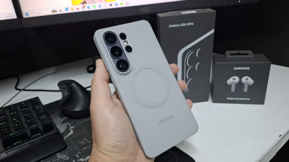 Mão segurando um smartphone Samsung Galaxy S26 Ultra cinza com anel MagSafe, ao lado das caixas do celular e do fone Galaxy Buds.