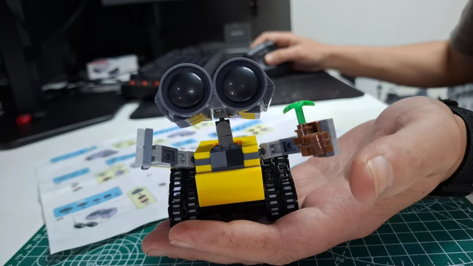 Mão segurando o mini robô WALL-E montado em frente ao manual sobre uma base de corte.