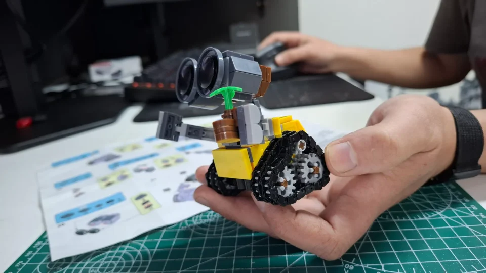 Mão segurando a base montada do robô WALL-E com as rodas e esteiras encaixadas sobre a mesa de trabalho.